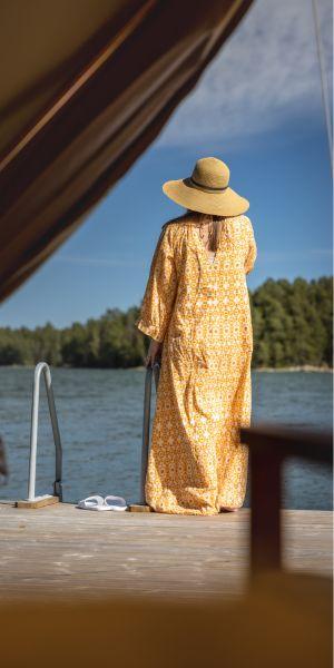 Kvinna på brygga med gul solklänning och solhatt. Blått vatten och blå himmel.