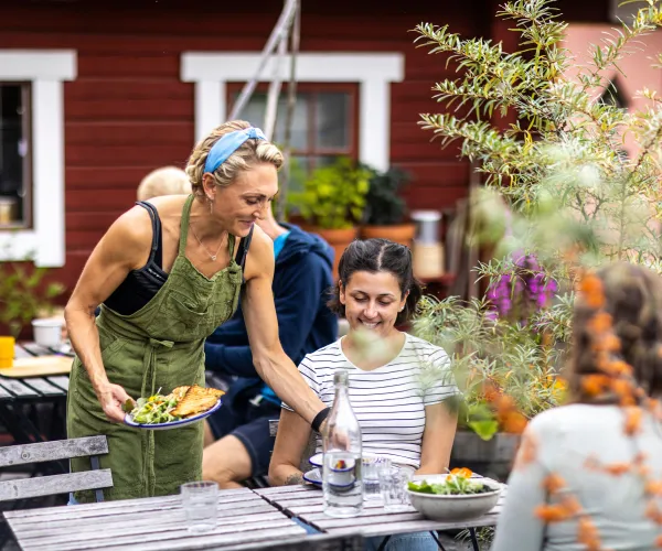 Uteservering framför röd stuga. Två gäster som blir serverade färgglad mat av servitris