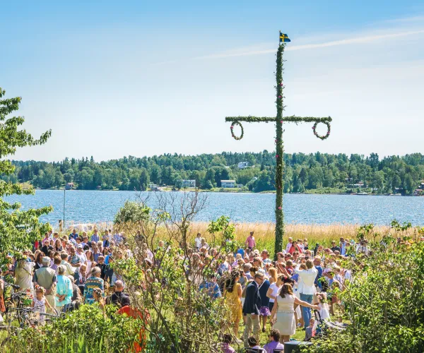 Midsommarfirande med folksamling kring midsommarstånd en solig dag med en blå vattenspegel i bakgrunden.