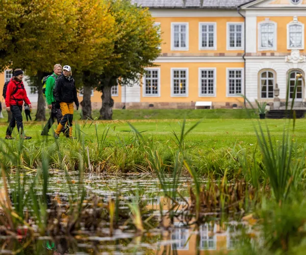 Tre höstklädda vandrare i en herrgårdsträdgård med herrgården i bakgrunden och en damm i förgrunden