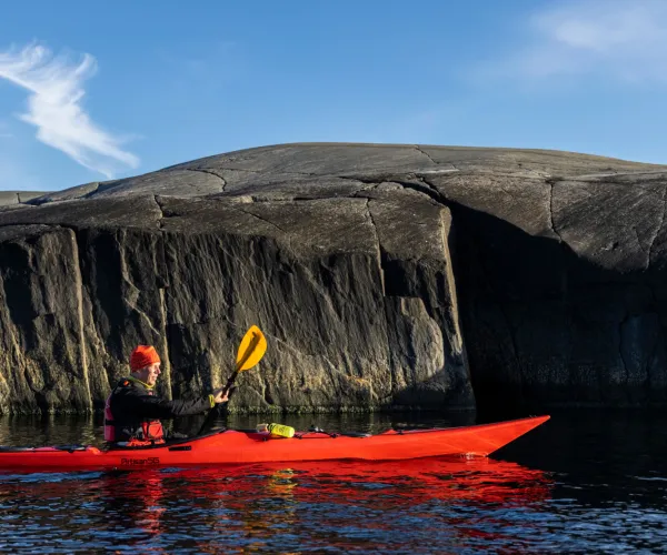 Paddlare i röd kajak i vattnet framför kal och brant klippa i ytterskärgården