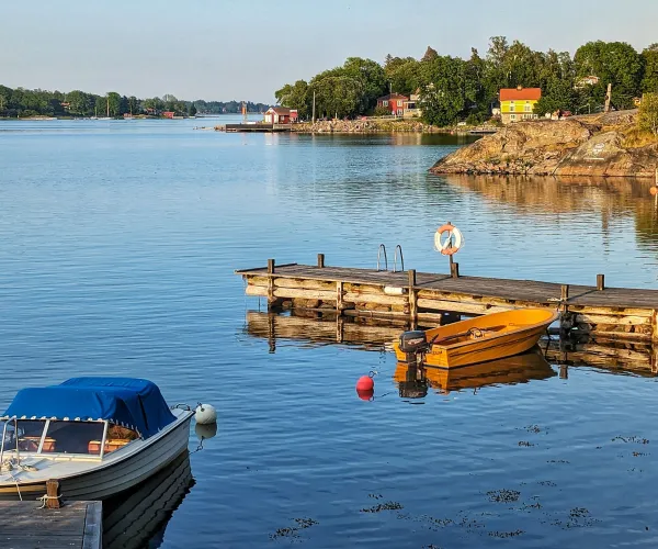Båt vid brygga i förgrunden. Blått hav och blå himmel. Gråa klippor, gul stuga och grön skog i bakgrunden